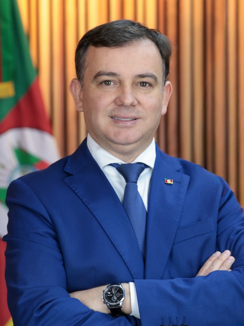 Homem de gravata e palet&oacute; azul pousa para foto oficial, &agrave; frente de uma bandeira do Rio Grande do Sul