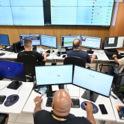 Sala de monitoramento com v&aacute;rias esta&ccedil;&otilde;es de trabalho alinhadas em mesas brancas. Cada esta&ccedil;&atilde;o possui dois monitores exibindo mapas, gr&aacute;ficos, listas de ocorr&ecirc;ncias e pain&eacute;is de controle digital.