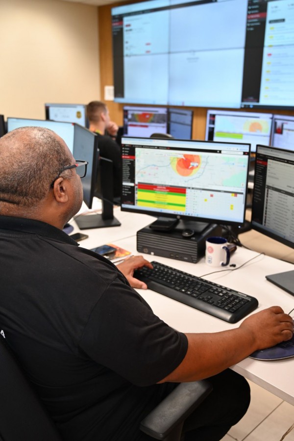 Vista lateral de uma estação de trabalho na central de monitoramento, com computador, teclado e mouse em uso. O monitor exibe mapa com áreas sinalizadas em cores, tabelas e indicadores operacionais.