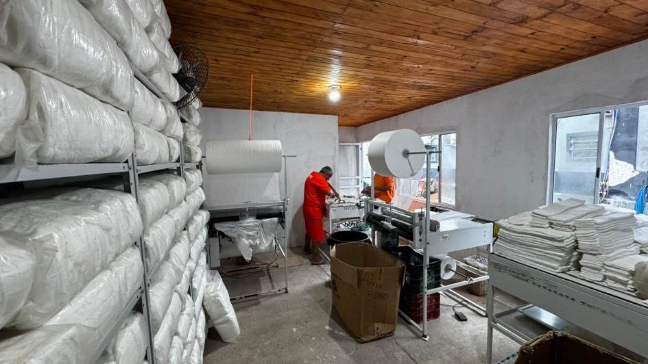 Ambiente interno de produ&ccedil;&atilde;o industrial, com estantes met&aacute;licas laterais carregadas com grandes volumes embalados em material pl&aacute;stico branco. No centro do espa&ccedil;o, h&aacute; m&aacute;quinas de processamento e embalagem, al&eacute;m de dois trabalhadores com uniformes cor de laranja. 