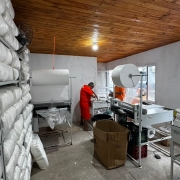 Ambiente interno de produ&ccedil;&atilde;o industrial, com estantes met&aacute;licas laterais carregadas com grandes volumes embalados em material pl&aacute;stico branco. No centro do espa&ccedil;o, h&aacute; m&aacute;quinas de processamento e embalagem, al&eacute;m de dois trabalhadores com uniformes cor de laranja. 