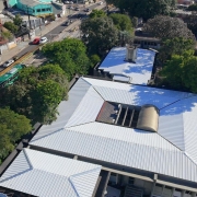 A fotografia colorida mostra um grande grupo de pessoas, composto por adultos e crian&ccedil;as, reunido em um p&aacute;tio escolar coberto para uma celebra&ccedil;&atilde;o coletiva.

Detalhes da cena:

O Grupo: O grupo &eacute; diversificado e ocupa quase todo o enquadramento. &Agrave; frente, crian&ccedil;as vestindo uniformes escolares (camisetas brancas ou verdes com detalhes em amarelo e azul) sorriem e acenam com as m&atilde;os para o alto. Atr&aacute;s delas, adultos &mdash; incluindo professores, gestores e possivelmente autoridades &mdash; tamb&eacute;m acenam e celebram.

Destaques: No centro do grupo, uma mulher segura um documento oficial emoldurado ou em uma pasta, indicando a formaliza&ccedil;&atilde;o de uma entrega ou projeto. Logo &agrave; frente, uma mulher agachada segura um microfone e sorri para a c&acirc;mera, sugerindo que ela coordenava o evento.

O Ambiente: O local &eacute; um p&aacute;tio coberto com p&eacute;-direito alto. A estrutura possui pilares e paredes pintadas em um tom de azul vibrante. O teto &eacute; forrado com placas brancas e possui lumin&aacute;rias embutidas. Ao fundo, as paredes s&atilde;o azuis e h&aacute; placas informativas fixadas.

Ilumina&ccedil;&atilde;o e Clima: A ilumina&ccedil;&atilde;o &eacute; natural e abundante, vinda das laterais abertas do p&aacute;tio. O clima da imagem &eacute; de grande entusiasmo, alegria e conquista compartilhada entre a comunidade escolar e os gestores presentes.

Composi&ccedil;&atilde;o: A foto &eacute; tirada de um &acirc;ngulo frontal e ligeiramente elevado, o que permite visualizar a profundidade do grupo e a express&atilde;o de felicidade de quase todos os participantes.