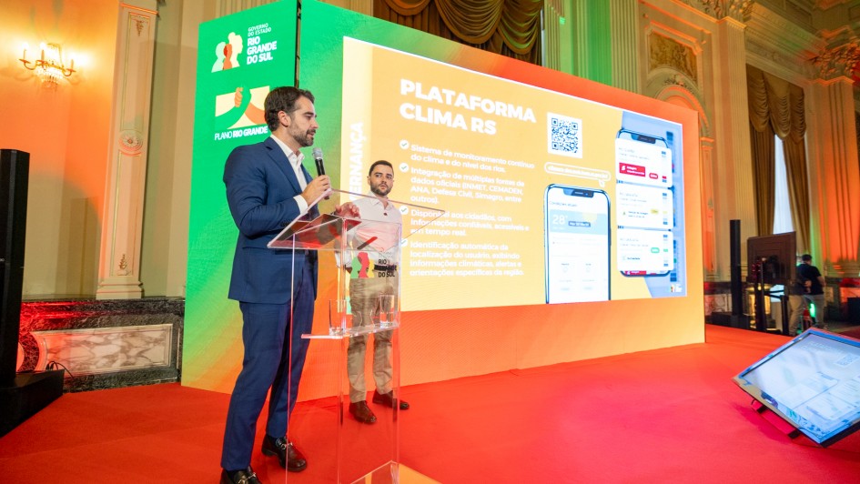 A fotografia colorida mostra o governador Eduardo Leite em um palco durante uma apresenta&ccedil;&atilde;o institucional em um sal&atilde;o nobre.

Detalhes da cena:

O orador: Eduardo Leite est&aacute; em p&eacute;, &agrave; esquerda, posicionado atr&aacute;s de um p&uacute;lpito de acr&iacute;lico transparente. Ele veste um terno azul, camisa branca e sapatos pretos. Segura um microfone com a m&atilde;o direita e gesticula com a m&atilde;o esquerda enquanto olha em dire&ccedil;&atilde;o &agrave; plateia (fora do quadro).

A apresenta&ccedil;&atilde;o: Atr&aacute;s dele, h&aacute; um grande tel&atilde;o de LED exibindo um slide com o t&iacute;tulo "PLATAFORMA CLIMA RS". O slide cont&eacute;m t&oacute;picos sobre o sistema de monitoramento, um QR Code no canto superior direito e ilustra&ccedil;&otilde;es de interfaces de aplicativos em smartphones.

O cen&aacute;rio: O evento ocorre em um local luxuoso, possivelmente o Pal&aacute;cio Piratini, com paredes ornamentadas, colunas cl&aacute;ssicas e ilumina&ccedil;&atilde;o c&ecirc;nica em tons de verde e &acirc;mbar. O ch&atilde;o do palco &eacute; coberto por um tapete vermelho vibrante. &Agrave; esquerda do tel&atilde;o, um painel verde exibe os logotipos do Governo do Estado do Rio Grande do Sul e do "Plano Rio Grande".

Outros elementos: Um segundo homem est&aacute; em p&eacute; ao fundo, atr&aacute;s do p&uacute;lpito, observando a apresenta&ccedil;&atilde;o. No canto inferior direito, uma pequena tela de retorno no ch&atilde;o reflete o conte&uacute;do do slide principal.

Composi&ccedil;&atilde;o e Ilumina&ccedil;&atilde;o: A imagem &eacute; capturada em um &acirc;ngulo diagonal, destacando a profundidade do palco e a grandiosidade do ambiente. A ilumina&ccedil;&atilde;o &eacute; focada no orador e no tel&atilde;o, criando um contraste n&iacute;tido com o restante do sal&atilde;o.