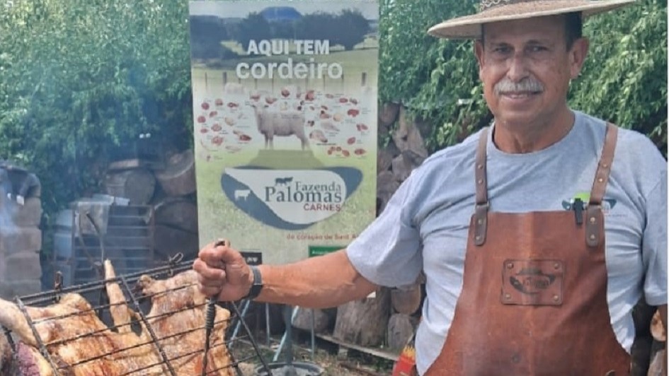 A imagem mostra uma cena ao ar livre de preparo de churrasco. Em primeiro plano, h&aacute; uma grande grelha inclinada com cortes de carne assando sobre o fogo. Ao lado da grelha, aparece um homem vestindo avental de couro e chap&eacute;u, segurando um utens&iacute;lio de ferro usado para manusear a estrutura. 