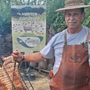 A imagem mostra uma cena ao ar livre de preparo de churrasco. Em primeiro plano, h&aacute; uma grande grelha inclinada com cortes de carne assando sobre o fogo. Ao lado da grelha, aparece um homem vestindo avental de couro e chap&eacute;u, segurando um utens&iacute;lio de ferro usado para manusear a estrutura. 