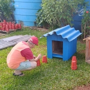 Uma pessoa est&aacute; em uma &aacute;rea externa gramada com pequenas estruturas de madeira em formato de casinhas, uma pintada de azul e outra em madeira natural. Ao redor, h&aacute; vasos pl&aacute;sticos vermelhos espalhados no ch&atilde;o e, ao fundo, pilhas organizadas de vasos semelhantes. 