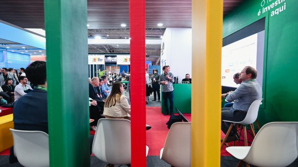 Foto mostra painel realizado no espa&ccedil;o do governo do Estado com painelistas e p&uacute;blico sentado. A cena &eacute; registrada atr&aacute;s de tr&ecirc;s pilares coloridos nas cores do Rio Grande do Sul, que est&atilde;o em primeiro plano. 