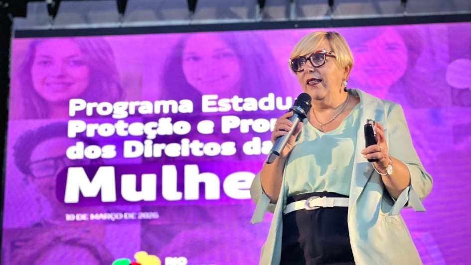 Uma mulher fala ao microfone em frente a um painel onde se l&ecirc; Programa Estadual de Prote&ccedil;&atilde;o e Promo&ccedil;&atilde;o dos Direitos das Mulheres.