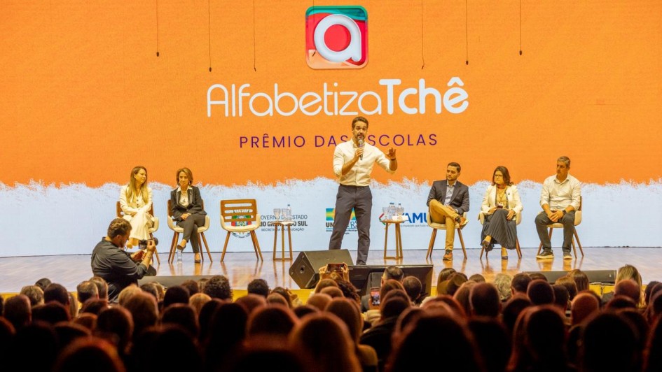 Vis&atilde;o ampla do palco durante evento, com o governador ao centro conduzindo a apresenta&ccedil;&atilde;o e outras pessoas sentadas em cadeiras alinhadas ao fundo. O painel LED exibe &ldquo;Alfabetiza Tch&ecirc; &ndash; Pr&ecirc;mio das Escolas&rdquo;, al&eacute;m de logotipos institucionais do governo estadual e parceiros. A plateia ocupa toda a &aacute;rea frontal do audit&oacute;rio.