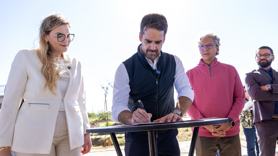 A fotografia colorida registra o ato solene de assinatura de um documento oficial pelo governador Eduardo Leite, em um ambiente externo sob luz solar intensa.

Detalhes da cena:

O ato da assinatura: No centro da imagem, o governador Eduardo Leite (homem branco, de barba e cabelos castanhos curtos) est&aacute; inclinado sobre uma mesa redonda alta de cor preta. Ele segura uma caneta com a m&atilde;o direita e assina um papel. Veste uma camisa social branca com as mangas dobradas e um colete preto por cima.

As autoridades ao redor:

&Agrave; esquerda, uma mulher loira de &oacute;culos com arma&ccedil;&atilde;o preta e blazer branco observa o documento com um sorriso discreto.

&Agrave; direita, um homem de cabelos grisalhos e &oacute;culos, vestindo um blus&atilde;o de l&atilde; rosa, observa o ato da assinatura.

Mais &agrave; direita, ao fundo, aparece um homem de terno cinza e barba, com os bra&ccedil;os cruzados.

O ambiente: O grupo est&aacute; sob uma estrutura de cobertura met&aacute;lica. Ao fundo, v&ecirc;-se um terreno aberto com vegeta&ccedil;&atilde;o rasteira e algumas &aacute;rvores jovens sob um c&eacute;u azul claro e limpo. A ilumina&ccedil;&atilde;o &eacute; natural e forte, vinda de tr&aacute;s e de cima, criando sombras marcadas.

Composi&ccedil;&atilde;o e enquadramento: A foto &eacute; um plano m&eacute;dio, capturando os participantes da cintura para cima. O foco est&aacute; no gesto da assinatura, simbolizando a formaliza&ccedil;&atilde;o de um investimento ou projeto p&uacute;blico.