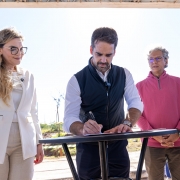 A fotografia colorida registra o ato solene de assinatura de um documento oficial pelo governador Eduardo Leite, em um ambiente externo sob luz solar intensa.

Detalhes da cena:

O ato da assinatura: No centro da imagem, o governador Eduardo Leite (homem branco, de barba e cabelos castanhos curtos) est&aacute; inclinado sobre uma mesa redonda alta de cor preta. Ele segura uma caneta com a m&atilde;o direita e assina um papel. Veste uma camisa social branca com as mangas dobradas e um colete preto por cima.

As autoridades ao redor:

&Agrave; esquerda, uma mulher loira de &oacute;culos com arma&ccedil;&atilde;o preta e blazer branco observa o documento com um sorriso discreto.

&Agrave; direita, um homem de cabelos grisalhos e &oacute;culos, vestindo um blus&atilde;o de l&atilde; rosa, observa o ato da assinatura.

Mais &agrave; direita, ao fundo, aparece um homem de terno cinza e barba, com os bra&ccedil;os cruzados.

O ambiente: O grupo est&aacute; sob uma estrutura de cobertura met&aacute;lica. Ao fundo, v&ecirc;-se um terreno aberto com vegeta&ccedil;&atilde;o rasteira e algumas &aacute;rvores jovens sob um c&eacute;u azul claro e limpo. A ilumina&ccedil;&atilde;o &eacute; natural e forte, vinda de tr&aacute;s e de cima, criando sombras marcadas.

Composi&ccedil;&atilde;o e enquadramento: A foto &eacute; um plano m&eacute;dio, capturando os participantes da cintura para cima. O foco est&aacute; no gesto da assinatura, simbolizando a formaliza&ccedil;&atilde;o de um investimento ou projeto p&uacute;blico.