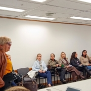 Aqui est&aacute; uma descri&ccedil;&atilde;o detalhada da imagem para fins de acessibilidade:

Descri&ccedil;&atilde;o da Imagem
A fotografia registra uma reuni&atilde;o institucional em um ambiente de escrit&oacute;rio formal. O enquadramento &eacute; em plano m&eacute;dio, capturando as participantes da esquerda para a direita.

Elementos Principais
Em primeiro plano (&agrave; esquerda): Uma mulher loira, de perfil, fala para o grupo. Ela usa &oacute;culos de arma&ccedil;&atilde;o escura, uma blusa mostarda e uma jaqueta de couro marrom. Ela parece liderar a conversa.

Ao fundo (centro e direita): Uma fileira de sete mulheres est&aacute; sentada em cadeiras pretas individuais, dispostas ao longo de uma parede branca. Elas ouvem atentamente a palestrante.

As vestimentas sugerem um clima frio e um tom profissional, incluindo blazers, cachec&oacute;is, su&eacute;teres e cal&ccedil;as sociais em tons de azul, marrom, cinza e preto.

Algumas seguram pastas ou cadernos no colo, indicando uma sess&atilde;o de trabalho ou planejamento.

O ambiente: A sala &eacute; iluminada por luzes fluorescentes embutidas em um forro de placas brancas. H&aacute; uma mesa branca longa que corta a parte inferior direita da imagem, onde se v&ecirc; parcialmente um tablet. No ch&atilde;o, bolsas e pastas das participantes est&atilde;o apoiadas perto das cadeiras.