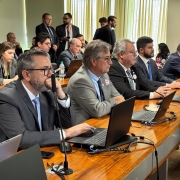 A imagem mostra uma sess&atilde;o institucional em uma sala de reuni&otilde;es ampla, com participantes sentados lado a lado em uma longa mesa de madeira. Cada posi&ccedil;&atilde;o possui notebook aberto, microfone individual, cabos organizados, celulares e garrafas de &aacute;gua. 