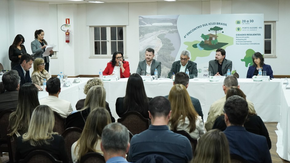 Aqui est&aacute; a descri&ccedil;&atilde;o detalhada da imagem para promover a acessibilidade:

Descri&ccedil;&atilde;o da Imagem
A fotografia registra a mesa principal do 4&ordm; Encontro Sul ICLEI Brasil, realizado em Porto Alegre. A cena &eacute; capturada de tr&aacute;s da plateia, mostrando a intera&ccedil;&atilde;o entre palestrantes e p&uacute;blico em um audit&oacute;rio.

Composi&ccedil;&atilde;o e Elementos
Mesa de Autoridades: Ao fundo, cinco integrantes comp&otilde;em a mesa principal, sentados atr&aacute;s de uma bancada longa coberta com uma toalha branca.

No centro-esquerda, uma mulher de palet&oacute; vermelho fala ao microfone.

Ao lado dela, tr&ecirc;s homens e uma mulher comp&otilde;em o restante da mesa, todos vestindo trajes formais (palet&oacute;s em tons de cinza e azul).

Sobre a mesa, h&aacute; garrafas de &aacute;gua mineral e pequenas placas de identifica&ccedil;&atilde;o para cada integrante.

Identifica&ccedil;&atilde;o do Evento: Atr&aacute;s da mesa, h&aacute; um painel informativo (banner) com o t&iacute;tulo: "4&ordm; ENCONTRO SUL ICLEI BRASIL". O painel destaca o tema "CIDADES RESILIENTES: Reconstru&ccedil;&atilde;o, Adapta&ccedil;&atilde;o e Financiamento P&oacute;s-Desastres Clim&aacute;ticos". H&aacute; tamb&eacute;m as datas (28 a 30 de abril) e a localiza&ccedil;&atilde;o (Porto Alegre - RS).

Plateia: Em primeiro plano, vemos as costas e as cabe&ccedil;as de diversas pessoas sentadas em cadeiras de madeira com estofado escuro. Elas est&atilde;o voltadas para a mesa principal, acompanhando a fala.

Ambiente: A sala possui ilumina&ccedil;&atilde;o clara, teto com lumin&aacute;rias embutidas e piso de madeira em padr&atilde;o chevron. No lado esquerdo, ao fundo, duas mulheres em p&eacute; organizam documentos pr&oacute;ximos a um extintor de inc&ecirc;ndio na parede.