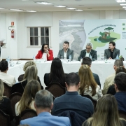 Aqui est&aacute; a descri&ccedil;&atilde;o detalhada da imagem para promover a acessibilidade:

Descri&ccedil;&atilde;o da Imagem
A fotografia registra a mesa principal do 4&ordm; Encontro Sul ICLEI Brasil, realizado em Porto Alegre. A cena &eacute; capturada de tr&aacute;s da plateia, mostrando a intera&ccedil;&atilde;o entre palestrantes e p&uacute;blico em um audit&oacute;rio.

Composi&ccedil;&atilde;o e Elementos
Mesa de Autoridades: Ao fundo, cinco integrantes comp&otilde;em a mesa principal, sentados atr&aacute;s de uma bancada longa coberta com uma toalha branca.

No centro-esquerda, uma mulher de palet&oacute; vermelho fala ao microfone.

Ao lado dela, tr&ecirc;s homens e uma mulher comp&otilde;em o restante da mesa, todos vestindo trajes formais (palet&oacute;s em tons de cinza e azul).

Sobre a mesa, h&aacute; garrafas de &aacute;gua mineral e pequenas placas de identifica&ccedil;&atilde;o para cada integrante.

Identifica&ccedil;&atilde;o do Evento: Atr&aacute;s da mesa, h&aacute; um painel informativo (banner) com o t&iacute;tulo: "4&ordm; ENCONTRO SUL ICLEI BRASIL". O painel destaca o tema "CIDADES RESILIENTES: Reconstru&ccedil;&atilde;o, Adapta&ccedil;&atilde;o e Financiamento P&oacute;s-Desastres Clim&aacute;ticos". H&aacute; tamb&eacute;m as datas (28 a 30 de abril) e a localiza&ccedil;&atilde;o (Porto Alegre - RS).

Plateia: Em primeiro plano, vemos as costas e as cabe&ccedil;as de diversas pessoas sentadas em cadeiras de madeira com estofado escuro. Elas est&atilde;o voltadas para a mesa principal, acompanhando a fala.

Ambiente: A sala possui ilumina&ccedil;&atilde;o clara, teto com lumin&aacute;rias embutidas e piso de madeira em padr&atilde;o chevron. No lado esquerdo, ao fundo, duas mulheres em p&eacute; organizam documentos pr&oacute;ximos a um extintor de inc&ecirc;ndio na parede.