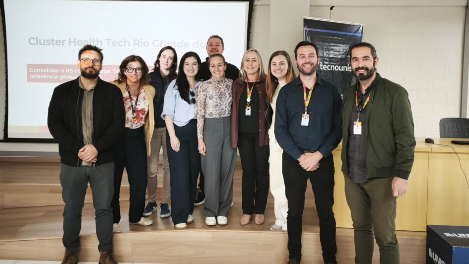 A imagem mostra um grupo reunido em um ambiente interno de apresenta&ccedil;&otilde;es, organizado para registro coletivo em frente a uma tela de proje&ccedil;&atilde;o. No fundo, &eacute; poss&iacute;vel ler o texto &ldquo;Cluster Health Tech Rio Grande do Sul&rdquo;.