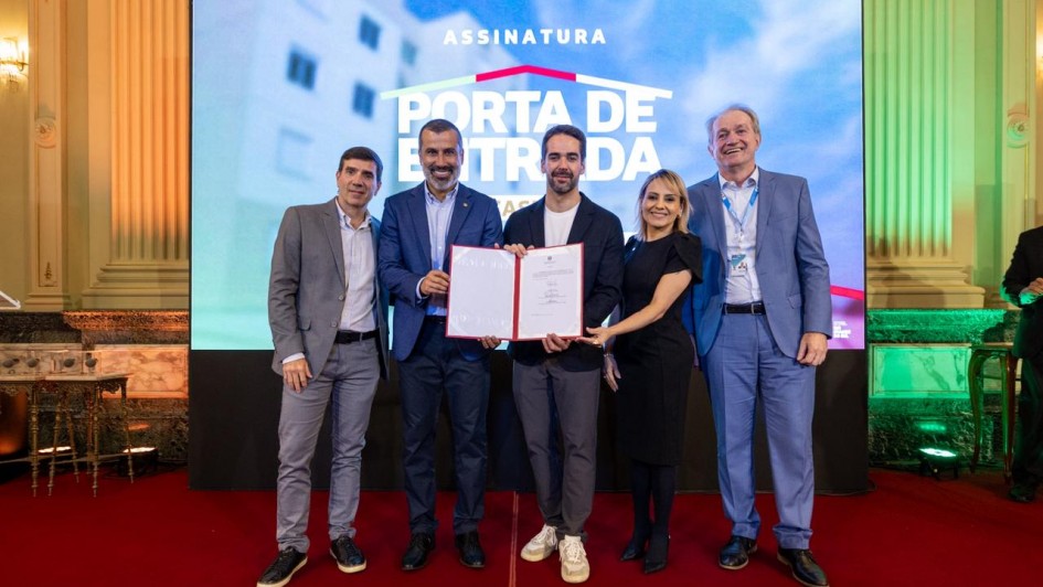 Grupo alinhado sobre o palco, posicionado diante de um painel LED com o texto &ldquo;ASSINATURA &ndash; PORTA DE ENTRADA &ndash; FASE 3&rdquo;. Ao centro, um documento aberto &eacute; exibido para registro oficial. 