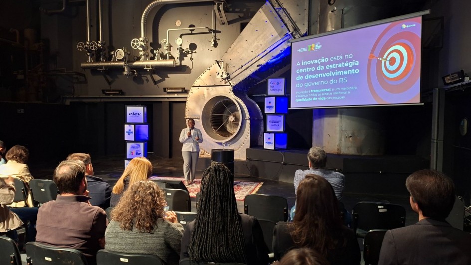 Aqui est&aacute; a descri&ccedil;&atilde;o detalhada da imagem para fins de acessibilidade:

Descri&ccedil;&atilde;o da Imagem
A fotografia captura uma palestra durante um evento preparat&oacute;rio para a Gramado Summit 2026. A cena ocorre em um ambiente com est&eacute;tica industrial moderna e ilumina&ccedil;&atilde;o dram&aacute;tica.

Elementos Principais
A Palestrante: No centro do palco, uma mulher negra veste um terno cinza claro. Ela est&aacute; de p&eacute; sobre um tapete persa, segurando um microfone e gesticulando enquanto fala para a plateia.

O Cen&aacute;rio: O fundo &eacute; composto por uma parede cinza escura com tubula&ccedil;&otilde;es industriais expostas, man&ocirc;metros e uma grande estrutura met&aacute;lica circular que se assemelha a uma turbina ou sistema de ventila&ccedil;&atilde;o.

A Apresenta&ccedil;&atilde;o: &Agrave; direita da palestrante, h&aacute; uma grande tela de proje&ccedil;&atilde;o exibindo um slide com fundo roxo. O texto diz: "A inova&ccedil;&atilde;o est&aacute; no centro da estrat&eacute;gia de desenvolvimento do governo do RS". O slide tamb&eacute;m inclui o desenho de um alvo com um dardo no centro e o logotipo do Governo do Estado do Rio Grande do Sul.

Elementos de Marca: Pr&oacute;ximo &agrave; palestrante, h&aacute; cubos iluminados empilhados exibindo logotipos de parceiros e eventos, como Gramado Summit, Sebrae, Google for Startups e RS Inova&ccedil;&atilde;o.

A Plateia: Em primeiro plano, vemos a parte de tr&aacute;s das cabe&ccedil;as e ombros de v&aacute;rias pessoas sentadas em cadeiras pretas, focadas na apresenta&ccedil;&atilde;o. A ilumina&ccedil;&atilde;o do audit&oacute;rio &eacute; reduzida, destacando apenas o palco e a tela.