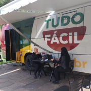 A fotografia mostra a Unidade M&oacute;vel do Tudo F&aacute;cil estacionada em um local p&uacute;blico ao ar livre, em Farroupilha. A cena foca no atendimento ao cidad&atilde;o realizado na parte externa do ve&iacute;culo.

Elementos Principais
O Ve&iacute;culo: &Agrave; esquerda e ao centro, v&ecirc;-se a lateral de um &ocirc;nibus adaptado. Ele &eacute; predominantemente branco com detalhes em verde e amarelo na parte frontal. Na lateral, h&aacute; um grande logotipo com as palavras "TUDO" em verde e "F&Aacute;CIL" em branco dentro de uma forma geom&eacute;trica vermelha. O &ocirc;nibus possui um adesivo com o s&iacute;mbolo internacional de acessibilidade pr&oacute;ximo &agrave; porta.

O Atendimento: Uma mesa preta dobr&aacute;vel est&aacute; montada ao lado do &ocirc;nibus, sob a prote&ccedil;&atilde;o de um toldo branco estendido a partir da lateral do ve&iacute;culo.

Atendente: &Agrave; direita, uma mulher de cabelos longos e pretos, vestida de preto, est&aacute; sentada operando um notebook.

Cidad&atilde;o: &Agrave; esquerda da mesa, um homem jovem, careca e tamb&eacute;m vestido de preto, est&aacute; sentado de frente para a atendente, aguardando ou acompanhando o servi&ccedil;o.

O Ambiente: O ch&atilde;o &eacute; de lajes de pedra cinza irregulares. O local parece ser um parque ou pra&ccedil;a, com gramado vis&iacute;vel ao fundo, &agrave; esquerda. A luz do dia sugere um ambiente externo bem iluminado.

Resumo visual: A imagem ilustra a presta&ccedil;&atilde;o de servi&ccedil;os p&uacute;blicos itinerantes. A estrutura m&oacute;vel do Tudo F&aacute;cil permite que o governo leve atendimento de documenta&ccedil;&atilde;o e servi&ccedil;os digitais diretamente &agrave;s comunidades, como exemplificado pelo atendimento personalizado na mesa externa.