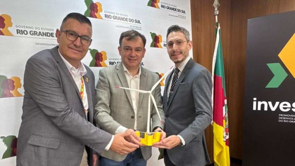 A fotografia registra um encontro institucional entre representantes do Governo do Estado e executivos, celebrando um investimento em energia renov&aacute;vel. Tr&ecirc;s homens est&atilde;o em p&eacute;, sorrindo para a c&acirc;mera, posicionados &agrave; frente de um painel oficial.

Elementos Principais
Os Participantes: * &Agrave; esquerda: Um homem de &oacute;culos e palet&oacute; cinza sobre camisa branca.

Ao centro: Um homem com palet&oacute; cinza claro xadrez e camisa branca sem gravata.

&Agrave; direita: Um homem de &oacute;culos, palet&oacute; cinza escuro, camisa branca e gravata com estampa discreta.

O Objeto Central: Os tr&ecirc;s homens seguram juntos uma maquete de uma turbina e&oacute;lica flutuante. A miniatura tem uma base amarela (que representa a plataforma flutuante) e uma torre branca com tr&ecirc;s p&aacute;s de h&eacute;lice.

O Plano de Fundo: * Logo atr&aacute;s deles, h&aacute; um painel branco repetindo o logotipo oficial do Governo do Estado do Rio Grande do Sul e da Secretaria de Desenvolvimento Econ&ocirc;mico.

&Agrave; direita, v&ecirc;-se a bandeira do Rio Grande do Sul em um mastro e um totem preto com a palavra "Invest" e o logotipo da ag&ecirc;ncia de fomento estadual.

Ambiente
O cen&aacute;rio &eacute; um gabinete ou sala de recep&ccedil;&atilde;o oficial com teto de placas brancas e divis&oacute;rias de madeira, indicando um ambiente governamental formal.

Resumo visual: A imagem simboliza a chegada de um grande investimento tecnol&oacute;gico ao estado. O foco na maquete da turbina e&oacute;lica flutuante destaca o pioneirismo do projeto de energia limpa mencionado no t&iacute;tulo.