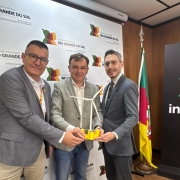 A fotografia registra um encontro institucional entre representantes do Governo do Estado e executivos, celebrando um investimento em energia renov&aacute;vel. Tr&ecirc;s homens est&atilde;o em p&eacute;, sorrindo para a c&acirc;mera, posicionados &agrave; frente de um painel oficial.

Elementos Principais
Os Participantes: * &Agrave; esquerda: Um homem de &oacute;culos e palet&oacute; cinza sobre camisa branca.

Ao centro: Um homem com palet&oacute; cinza claro xadrez e camisa branca sem gravata.

&Agrave; direita: Um homem de &oacute;culos, palet&oacute; cinza escuro, camisa branca e gravata com estampa discreta.

O Objeto Central: Os tr&ecirc;s homens seguram juntos uma maquete de uma turbina e&oacute;lica flutuante. A miniatura tem uma base amarela (que representa a plataforma flutuante) e uma torre branca com tr&ecirc;s p&aacute;s de h&eacute;lice.

O Plano de Fundo: * Logo atr&aacute;s deles, h&aacute; um painel branco repetindo o logotipo oficial do Governo do Estado do Rio Grande do Sul e da Secretaria de Desenvolvimento Econ&ocirc;mico.

&Agrave; direita, v&ecirc;-se a bandeira do Rio Grande do Sul em um mastro e um totem preto com a palavra "Invest" e o logotipo da ag&ecirc;ncia de fomento estadual.

Ambiente
O cen&aacute;rio &eacute; um gabinete ou sala de recep&ccedil;&atilde;o oficial com teto de placas brancas e divis&oacute;rias de madeira, indicando um ambiente governamental formal.

Resumo visual: A imagem simboliza a chegada de um grande investimento tecnol&oacute;gico ao estado. O foco na maquete da turbina e&oacute;lica flutuante destaca o pioneirismo do projeto de energia limpa mencionado no t&iacute;tulo.