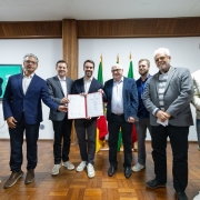 Registro coletivo em ambiente interno, com um grupo alinhado diante de bandeiras oficiais posicionadas ao fundo. No centro, um documento aberto &eacute; exibido, indicando ato formal de assinatura ou apresenta&ccedil;&atilde;o.