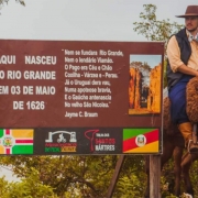 Aqui est&aacute; a descri&ccedil;&atilde;o detalhada da imagem para fins de acessibilidade:

Descri&ccedil;&atilde;o da Imagem
A fotografia mostra um marco hist&oacute;rico ao ar livre, destacando uma placa informativa e pessoas vestidas com trajes t&iacute;picos ga&uacute;chos montadas em mulas. A cena evoca a preserva&ccedil;&atilde;o da cultura e hist&oacute;ria regional.

Elementos Principais
A Placa Informativa: No centro da imagem, h&aacute; uma grande placa retangular marrom sustentada por postes de madeira.

Texto &agrave; esquerda: Em letras brancas e mai&uacute;sculas, l&ecirc;-se: "AQUI NASCEU O RIO GRANDE EM 03 DE MAIO DE 1626".

Texto ao centro: Um poema de Jayme C. Braun impresso em branco, que exalta a hist&oacute;ria do Rio Grande e de S&atilde;o Nicolau.

Lado direito da placa: Uma foto vertical de ru&iacute;nas hist&oacute;ricas (provavelmente das Miss&otilde;es).

Rodap&eacute; da placa: Exibe logotipos e bandeiras, incluindo a bandeira do Rio Grande do Sul, a bandeira de S&atilde;o Nicolau e s&iacute;mbolos de projetos tur&iacute;sticos como "Missioneiros do Pedal" e "Trilha dos Santos M&aacute;rtires".

Os Cavaleiros: &Agrave; direita da placa, um homem e uma mulher aparecem montados em mulas de pelagem escura.

Ambos vestem indument&aacute;ria tradicional ga&uacute;cha: chap&eacute;us de aba larga e copa baixa (barbicacho), len&ccedil;os no pesco&ccedil;o e coletes.

O homem, em primeiro plano, usa bombacha azul e botas de couro. A sela da mula est&aacute; coberta por um pelego de l&atilde; marrom.

O Ambiente: O cen&aacute;rio &eacute; rural, com vegeta&ccedil;&atilde;o verde baixa ao redor da placa e &aacute;rvores ao fundo. O c&eacute;u est&aacute; nublado, conferindo uma ilumina&ccedil;&atilde;o suave e difusa &agrave; imagem. &Agrave; esquerda, ao fundo, percebe-se uma extens&atilde;o de &aacute;gua (possivelmente um rio) e uma cerca de arame farpado.

Resumo visual: A imagem celebra a funda&ccedil;&atilde;o hist&oacute;rica do Rio Grande do Sul em S&atilde;o Nicolau, unindo o registro oficial do marco hist&oacute;rico &agrave; figura viva do ga&uacute;cho contempor&acirc;neo que preserva suas tradi&ccedil;&otilde;es.