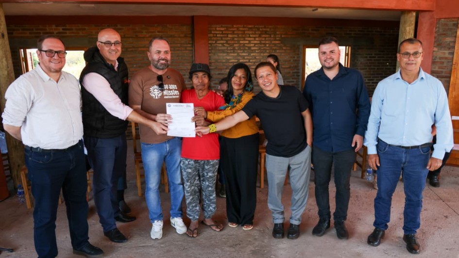 Registro coletivo em ambiente interno r&uacute;stico, com paredes de tijolo aparente e piso simples. Um grupo est&aacute; alinhado para foto, e ao centro um documento &eacute; exibido.