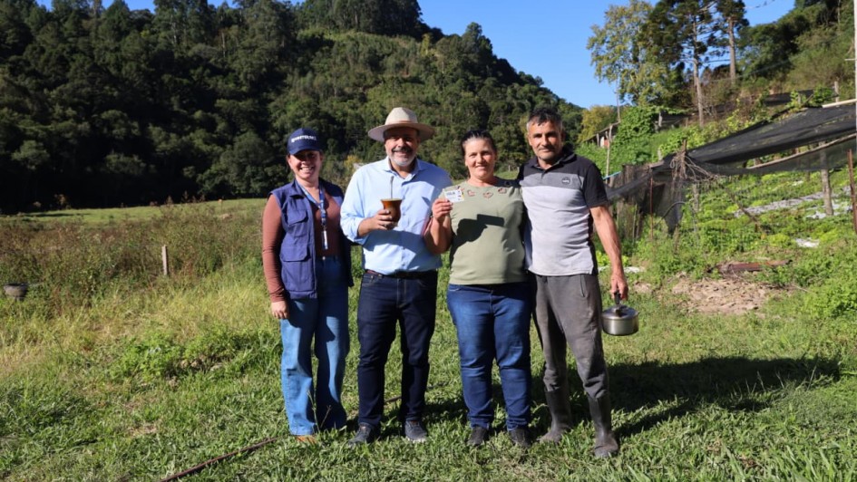A imagem mostra um registro em &aacute;rea rural, com quatro pessoas posicionadas lado a lado em um campo gramado. Ao fundo, h&aacute; vegeta&ccedil;&atilde;o densa, &aacute;rvores altas e um relevo coberto por mata, sob c&eacute;u azul e ilumina&ccedil;&atilde;o natural intensa. Uma das pessoas segura um cart&atilde;o pl&aacute;stico identificado como Cart&atilde;o Cidad&atilde;o.