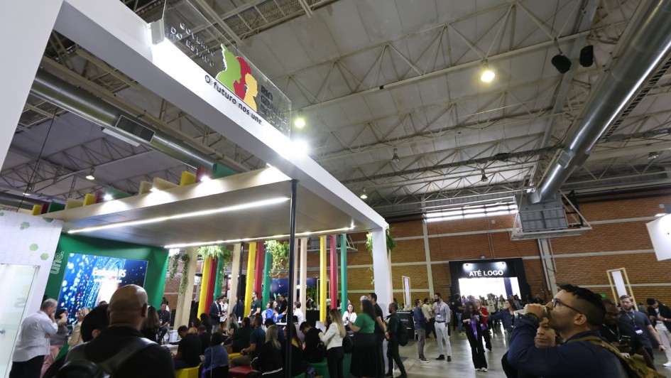 A imagem mostra um estande institucional em um grande pavilh&atilde;o de eventos, com estrutura moderna em tons claros e ilumina&ccedil;&atilde;o integrada. No topo do estande h&aacute; um painel transparente com a marca do Governo do Estado do Rio Grande do Sul e a frase &ldquo;Futuro nos une&rdquo;. Ao redor, h&aacute; intensa circula&ccedil;&atilde;o de visitantes pelos corredores do pavilh&atilde;o.
