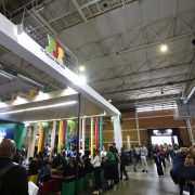 A imagem mostra um estande institucional em um grande pavilh&atilde;o de eventos, com estrutura moderna em tons claros e ilumina&ccedil;&atilde;o integrada. No topo do estande h&aacute; um painel transparente com a marca do Governo do Estado do Rio Grande do Sul e a frase &ldquo;Futuro nos une&rdquo;. Ao redor, h&aacute; intensa circula&ccedil;&atilde;o de visitantes pelos corredores do pavilh&atilde;o.