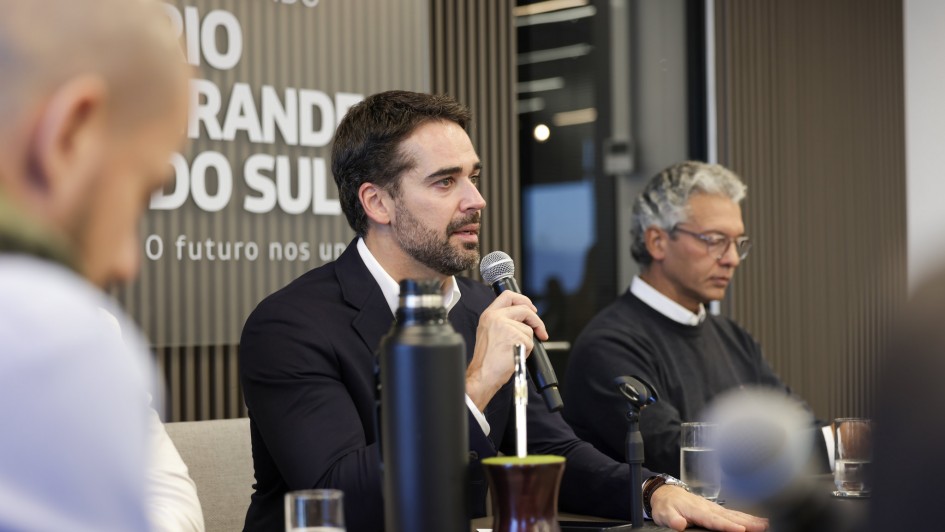 Aqui est&aacute; a descri&ccedil;&atilde;o detalhada da imagem para fins de acessibilidade:

Descri&ccedil;&atilde;o da Imagem
A fotografia registra uma coletiva de imprensa ou pronunciamento oficial. O foco principal est&aacute; no Governador do Rio Grande do Sul, Eduardo Leite, que fala ao microfone.

Elementos Principais
Eduardo Leite (Centro): O governador est&aacute; de perfil para a direita, segurando um microfone pr&oacute;ximo &agrave; boca. Ele possui barba aparada, cabelo castanho curto e veste um palet&oacute; azul-marinho sobre uma camisa branca de gola aberta. Sua express&atilde;o &eacute; s&eacute;ria e concentrada.

Outro Participante (Direita): Ao lado dele, um homem de cabelos grisalhos e &oacute;culos, vestindo um su&eacute;ter escuro sobre camisa branca, observa o evento com uma postura atenta e reservada.

Elementos Culturais sobre a Mesa: &Agrave; frente do governador, sobre uma mesa escura, destacam-se itens t&iacute;picos da cultura ga&uacute;cha: um cuia de mate com erva-verde e uma bomba de metal, al&eacute;m de uma garrafa t&eacute;rmica preta alta. Tamb&eacute;m h&aacute; copos de &aacute;gua sobre a mesa.

Plano de Fundo: Atr&aacute;s dos oradores, h&aacute; um painel decorativo com ripas verticais de madeira. No lado esquerdo do painel, l&ecirc;-se parte da identidade visual do governo: "GOVERNO DO ESTADO RIO GRANDE DO SUL" e o slogan "O futuro nos une".

Composi&ccedil;&atilde;o: A imagem utiliza uma profundidade de campo reduzida, deixando uma pessoa em primeiro plano (&agrave; esquerda, de costas) e o fundo levemente desfocados para manter o foco total nas autoridades e no pronunciamento.

Resumo visual: A imagem retrata um momento de lideran&ccedil;a e comunica&ccedil;&atilde;o governamental, contextualizado por s&iacute;mbolos da tradi&ccedil;&atilde;o ga&uacute;cha, sugerindo um an&uacute;ncio oficial ou reuni&atilde;o estrat&eacute;gica do Governo do Estado.