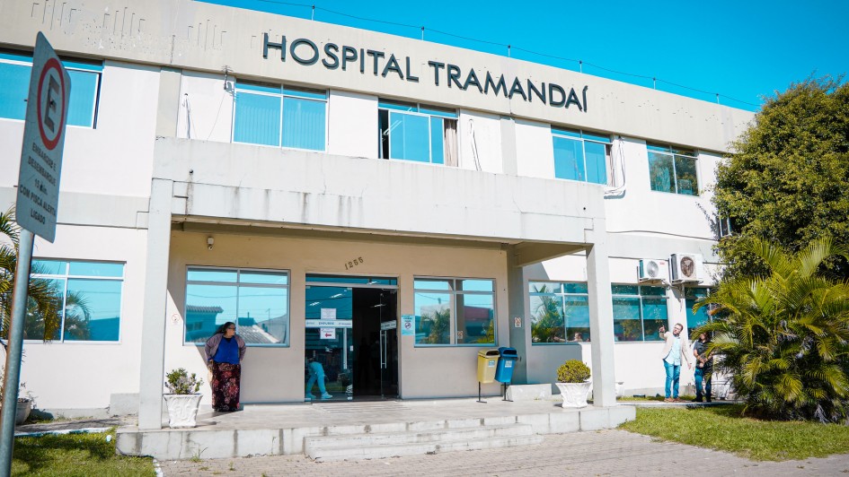 A fotografia apresenta a fachada principal do Hospital Tramanda&iacute;, capturada sob luz solar intensa e c&eacute;u azul sem nuvens. O edif&iacute;cio possui dois pavimentos e uma arquitetura funcional em tons claros.

Elementos Principais
Fachada do Edif&iacute;cio: O pr&eacute;dio &eacute; pintado de branco ou bege claro. No topo da fachada frontal, destaca-se o letreiro em letras pretas e mai&uacute;sculas: "HOSPITAL TRAMANDA&Iacute;".

Entrada: A entrada principal &eacute; protegida por uma marquise de concreto sustentada por pilares. A porta de vidro central &eacute; ladeada por grandes janelas que refletem o c&eacute;u azul. Acima da porta, v&ecirc;-se o n&uacute;mero 1255.

Pessoas e Atividade:

&Agrave; esquerda da entrada, uma mulher vestindo uma saia longa estampada e um casaco azul aguarda em p&eacute; na cal&ccedil;ada.

&Agrave; direita, no gramado, dois homens conversam; um deles veste camisa azul e cal&ccedil;a jeans, apontando em dire&ccedil;&atilde;o ao pr&eacute;dio.

Detalhes Adicionais:

Sinaliza&ccedil;&atilde;o: No canto esquerdo, em primeiro plano, h&aacute; uma placa de tr&acirc;nsito de regulamenta&ccedil;&atilde;o (proibido estacionar, com exce&ccedil;&atilde;o para embarque e desembarque r&aacute;pido).

Vegeta&ccedil;&atilde;o: O hospital &eacute; ladeado por &aacute;reas gramadas verdes, com palmeiras e arbustos decorativos em vasos brancos na entrada.

Infraestrutura: V&ecirc;-se aparelhos de ar-condicionado instalados na parede externa do segundo andar e lixeiras de coleta seletiva (azul e amarela) pr&oacute;ximas &agrave; entrada.

Resumo visual: A imagem retrata a unidade de sa&uacute;de em um dia claro, transmitindo um aspecto de organiza&ccedil;&atilde;o e atendimento p&uacute;blico regular na regi&atilde;o litor&acirc;nea.