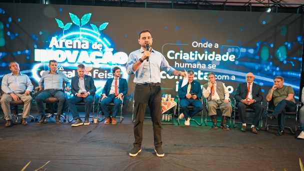 Homem discursa com microfone em um palco, atr&aacute;s dele v&aacute;rios outros homens est&atilde;o sentados e mais atr&aacute;s um grande banner com dizeres "Arena de inova&ccedil;&atilde;o"