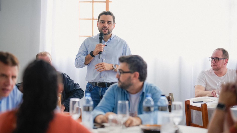 Homem fala ao microfone em um almo&ccedil;o, observado por v&aacute;rias pessoas sentadas em um ambiente de almo