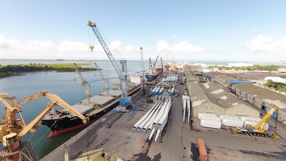A fotografia a&eacute;rea apresenta uma vista panor&acirc;mica de um porto comercial em plena atividade. O cen&aacute;rio &eacute; dominado por grandes infraestruturas log&iacute;sticas, navios de carga e componentes industriais sob um c&eacute;u claro com algumas nuvens esparsas.

Elementos Principais
Navios e Guindastes:

No lado esquerdo, um grande navio cargueiro de casco preto e detalhes em vermelho est&aacute; atracado no cais.

V&aacute;rios guindastes de grande porte, em cores como laranja, azul e amarelo, est&atilde;o posicionados ao longo do porto para a movimenta&ccedil;&atilde;o de cargas pesadas.

Carga em Destaque:

No centro do p&aacute;tio portu&aacute;rio, destacam-se v&aacute;rias p&aacute;s de turbinas e&oacute;licas brancas e longas, organizadas horizontalmente no ch&atilde;o, aguardando transporte ou embarque.

Pr&oacute;ximo a elas, h&aacute; outros componentes cil&iacute;ndricos azuis e grandes caixas brancas protegidas.

Armaz&eacute;ns e Estruturas:

&Agrave; direita, estende-se uma fileira de grandes galp&otilde;es ou armaz&eacute;ns industriais com telhados cinzas de duas &aacute;guas.

Containers de diversas cores est&atilde;o espalhados pelas &aacute;reas de p&aacute;tio.

Geografia:

O porto est&aacute; situado &agrave; beira de um corpo d'&aacute;gua esverdeado.

Ao fundo, no lado esquerdo, v&ecirc;-se uma &aacute;rea de vegeta&ccedil;&atilde;o rasteira e densa (provavelmente mangue ou &aacute;rea de preserva&ccedil;&atilde;o), separada por canais de &aacute;gua. No horizonte, a linha da costa se estende sob o c&eacute;u azul.

Resumo visual: A imagem captura a complexidade e a escala de uma opera&ccedil;&atilde;o portu&aacute;ria moderna, evidenciando o papel do porto na log&iacute;stica de energia renov&aacute;vel (componentes e&oacute;licos) e no com&eacute;rcio mar&iacute;timo global.