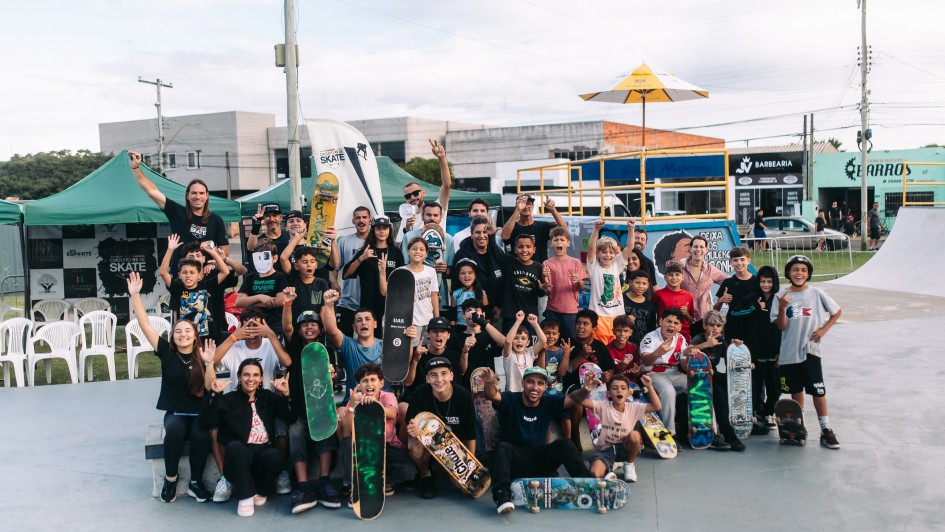 A imagem mostra um registro coletivo ao ar livre em pista de skate, com um grupo numeroso reunido para foto em frente a obst&aacute;culos do espa&ccedil;o esportivo. No primeiro plano, est&atilde;o skates apoiados no ch&atilde;o e sendo segurados, enquanto participantes permanecem em diferentes n&iacute;veis, alguns sentados e outros em p&eacute;.