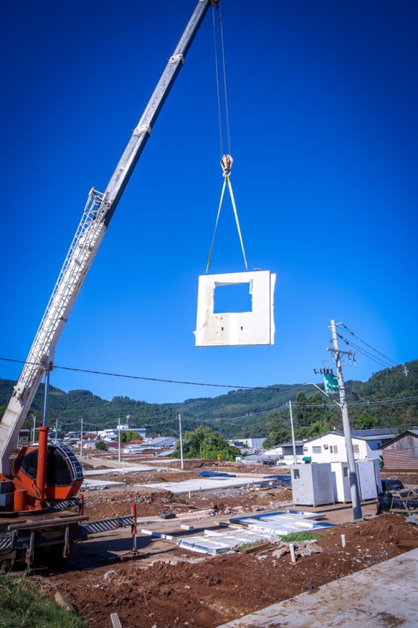 Foto mostra painel de concreto sendo içado por guindaste na obra das casas no loteamento em Muçum.