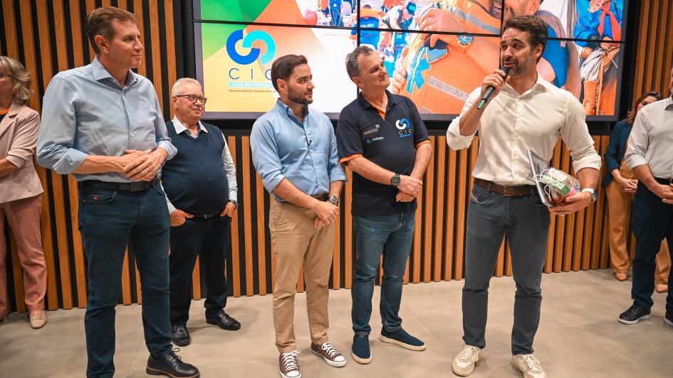 Grupo alinhado em palco durante evento institucional, com painel digital ao fundo exibindo logotipo CIC Vale do Taquari e imagens de a&ccedil;&otilde;es sociais. O governador fala ao microfone segurando um objeto simb&oacute;lico, enquanto demais pessoas permanecem posicionadas ao lado.