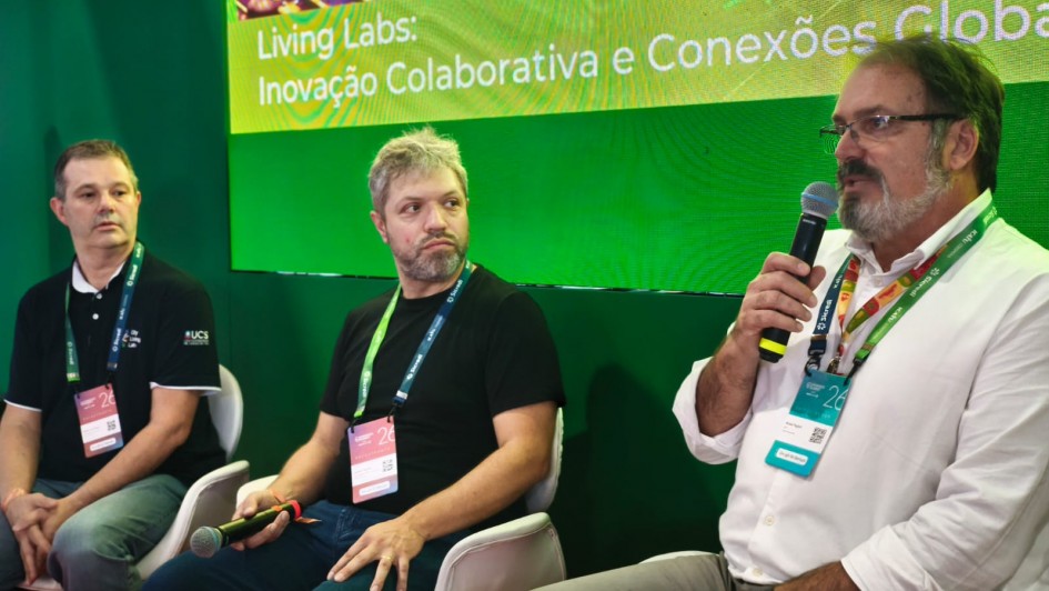 Tr&ecirc;s homens est&atilde;o sentados em um palco, com um painel ao fundo, onde se l&ecirc; "Living Labs: inova&ccedil;&atilde;o colaborativa e conex&otilde;es globais". 