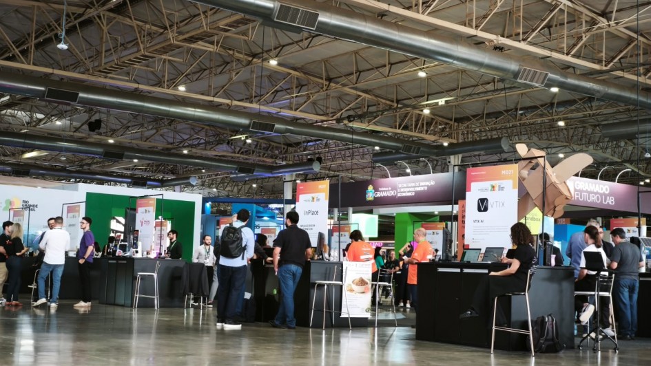 Esta &eacute; uma fotografia de um amplo pavilh&atilde;o de eventos durante uma feira de inova&ccedil;&atilde;o e tecnologia, focada na &aacute;rea de startups.

Estrutura do Ambiente
Teto Industrial: O teto &eacute; alto, com estrutura met&aacute;lica aparente (treli&ccedil;as), grandes tubula&ccedil;&otilde;es de ventila&ccedil;&atilde;o prateadas e ilumina&ccedil;&atilde;o industrial distribu&iacute;da por toda a extens&atilde;o.

Piso: O ch&atilde;o &eacute; de concreto polido cinza, que reflete suavemente a luz e o movimento das pessoas.

Estandes: No plano m&eacute;dio, veem-se diversos balc&otilde;es de atendimento pretos, organizados de forma linear. Cada balc&atilde;o representa uma empresa ou startup diferente, identificadas por banners verticais brancos com logotipos e textos coloridos.

Elementos em Destaque
Identifica&ccedil;&otilde;es: &Eacute; poss&iacute;vel ler nomes como "InPlace", "VTIX" e refer&ecirc;ncias institucionais como "Prefeitura de Gramado" e "Gramado Futuro Lab".

Pessoas: Grupos de pessoas circulam pelo espa&ccedil;o ou est&atilde;o paradas junto aos balc&otilde;es conversando. O vestu&aacute;rio &eacute; predominantemente casual (camisetas, jeans e mochilas), t&iacute;pico de ambientes de tecnologia. Alguns vestem camisetas laranjas, possivelmente da organiza&ccedil;&atilde;o ou de uma startup espec&iacute;fica.

Decora&ccedil;&atilde;o Criativa: &Agrave; direita, destaca-se uma escultura de papel&atilde;o em formato de avi&atilde;o ou h&eacute;lice, montada sobre um dos estandes, refor&ccedil;ando a tem&aacute;tica de criatividade e "m&atilde;o na massa" (maker).

Composi&ccedil;&atilde;o e Atmosfera
A foto foi tirada de uma perspectiva ampla, mostrando a escala do evento. A ilumina&ccedil;&atilde;o &eacute; clara e uniforme, transmitindo um ambiente din&acirc;mico, profissional e focado em networking e neg&oacute;cios.