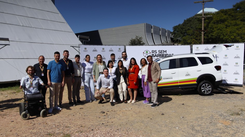 Esta imagem captura um momento oficial de celebra&ccedil;&atilde;o ou lan&ccedil;amento de um projeto governamental em um ambiente externo.

Pessoas e Composi&ccedil;&atilde;o
O Grupo: Aproximadamente 15 pessoas est&atilde;o reunidas e sorridentes para a fotografia. O grupo &eacute; diverso em g&ecirc;nero e idade, vestindo roupas casuais e formais de trabalho.

Inclus&atilde;o: No lado esquerdo da imagem, destaca-se um homem em uma cadeira de rodas motorizada, integrando o grupo.

Posicionamento: As pessoas est&atilde;o organizadas em duas fileiras (algumas em p&eacute;, outras levemente agachadas &agrave; frente) diante de um fundo composto por pain&eacute;is e um ve&iacute;culo.

Elementos de Identifica&ccedil;&atilde;o
Ve&iacute;culo Adaptado: &Agrave; direita, h&aacute; um SUV branco (modelo Chevrolet) com adesivos do Governo do Estado do Rio Grande do Sul e as cores da bandeira estadual. O porta-malas est&aacute; aberto, revelando uma rampa de acessibilidade instalada.

Pain&eacute;is: Ao fundo do grupo, pain&eacute;is brancos exibem repetidamente o logotipo do programa "RS SEM BARREIRAS - Acessibilidade para todos".

Cen&aacute;rio e Ilumina&ccedil;&atilde;o
Ambiente: A foto foi tirada em um terreno de terra e pedriscos, sob luz solar direta e forte, o que gera sombras marcadas no ch&atilde;o.

Arquitetura: Ao fundo, o cen&aacute;rio &eacute; composto por pr&eacute;dios de arquitetura moderna e imponente, com linhas geom&eacute;tricas e superf&iacute;cies cinzas e brancas, sob um c&eacute;u azul sem nuvens.

A imagem registra a formaliza&ccedil;&atilde;o de uma parceria para a cess&atilde;o de ve&iacute;culos adaptados, refor&ccedil;ando temas de mobilidade e pol&iacute;ticas p&uacute;blicas de inclus&atilde;o.