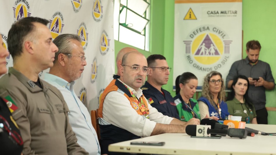 Esta fotografia registra uma coletiva de imprensa ou reuni&atilde;o t&eacute;cnica oficial, com foco em a&ccedil;&otilde;es de defesa civil e seguran&ccedil;a p&uacute;blica.

Elementos Principais e Pessoas
O Porta-voz: No centro da imagem, um homem calvo, usando &oacute;culos e um colete oficial da Defesa Civil (nas cores laranja, azul e branco), fala em dire&ccedil;&atilde;o aos microfones.

Composi&ccedil;&atilde;o da Mesa: Ele est&aacute; acompanhado por outros homens e mulheres sentados em uma longa mesa branca. &Agrave; sua esquerda, destaca-se um homem com uniforme militar (possivelmente da Brigada Militar ou Casa Militar), e &agrave; sua direita, um homem em um uniforme azul escuro. Outras pessoas, incluindo mulheres com coletes de identifica&ccedil;&atilde;o, completam a mesa.

Equipamentos de Imprensa: Sobre a mesa, est&atilde;o posicionados diversos microfones de ve&iacute;culos de comunica&ccedil;&atilde;o, sendo poss&iacute;vel identificar claramente o logotipo da RBS TV. H&aacute; tamb&eacute;m um smartphone e outros dispositivos de grava&ccedil;&atilde;o.

Cen&aacute;rio e Identifica&ccedil;&atilde;o Visual
Banners e Marcas: Ao fundo, o ambiente &eacute; caracterizado por elementos de identidade visual do Governo do Estado. H&aacute; um banner lateral repetindo os logos da "Casa Militar" e da "Defesa Civil - RS". Na parede ao fundo, um p&ocirc;ster maior exibe o bras&atilde;o da Defesa Civil do Rio Grande do Sul (um tri&acirc;ngulo com figuras humanas no centro).

Ambiente: A sala possui paredes em um tom de verde claro, com uma janela vis&iacute;vel ao fundo. A ilumina&ccedil;&atilde;o &eacute; de interior, focada nos participantes da mesa.

Atmosfera
A imagem transmite um clima de seriedade e coordena&ccedil;&atilde;o institucional, t&iacute;pico de atualiza&ccedil;&otilde;es de emerg&ecirc;ncia ou an&uacute;ncios de planos de conting&ecirc;ncia governamentais.