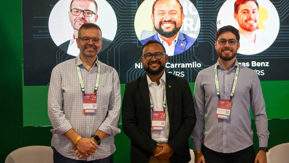 Esta fotografia registra um momento de apresenta&ccedil;&atilde;o tecnol&oacute;gica em um evento corporativo ou governamental focado em inova&ccedil;&atilde;o.

Pessoas e Composi&ccedil;&atilde;o
Os Palestrantes: Tr&ecirc;s homens est&atilde;o em p&eacute;, lado a lado, sorrindo para a c&acirc;mera. O homem &agrave; esquerda veste uma camisa de mangas longas com padronagem xadrez mi&uacute;da. O homem ao centro veste uma camisa branca sob um palet&oacute; preto. O homem &agrave; direita veste uma camisa social lil&aacute;s clara.

Identifica&ccedil;&atilde;o: Todos utilizam crach&aacute;s de identifica&ccedil;&atilde;o pendurados no pesco&ccedil;o, onde &eacute; poss&iacute;vel ler a palavra "PALESTRANTE" e o n&uacute;mero "26".

Elementos Visuais e Cen&aacute;rio
Tela de Fundo: Atr&aacute;s do grupo, h&aacute; um grande painel digital de cor verde vibrante. A tela exibe um design que remete a circuitos eletr&ocirc;nicos e processadores. Nela, est&atilde;o projetados tr&ecirc;s c&iacute;rculos contendo as fotos de rosto de cada um dos palestrantes, acompanhadas de seus respectivos nomes, como "Nilson Carramilo" e "Lucas Benz".

Mobili&aacute;rio: Na parte inferior da imagem, aparecem os encostos curvos de tr&ecirc;s poltronas brancas, sugerindo que o grupo est&aacute; em um palco ou &aacute;rea de debates.

Contexto
O t&iacute;tulo do arquivo indica que a imagem se refere &agrave; apresenta&ccedil;&atilde;o das iniciativas GurIA e Aurora, descritas como estrat&eacute;gias para a gest&atilde;o p&uacute;blica.