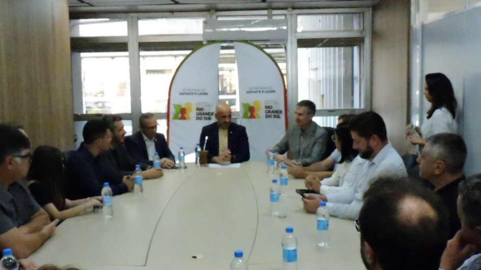           Esta fotografia retrata uma reuni&atilde;o oficial de trabalho realizada em uma sala de confer&ecirc;ncias institucional.

Elementos Centrais e Pessoas
A Reuni&atilde;o: Cerca de doze pessoas est&atilde;o sentadas ao redor de uma grande mesa oval de cor clara. Os participantes, homens e mulheres, vestem trajes que variam entre o social e o casual profissional.

Destaque: Na cabeceira da mesa, ao fundo, um homem fala aos presentes enquanto segura uma cuia de mate, um elemento cultural caracter&iacute;stico. &Agrave; frente de quase todos os participantes, h&aacute; garrafas de &aacute;gua individuais sobre a mesa.

Cen&aacute;rio e Identifica&ccedil;&atilde;o
Banner Institucional: Ao fundo, atrav&eacute;s de uma divis&oacute;ria de vidro ou porta, &eacute; poss&iacute;vel ver um banner branco com o bras&atilde;o do Governo do Estado e o nome "Secretaria do Esporte e Lazer".

Ambiente: A sala possui ilumina&ccedil;&atilde;o clara de escrit&oacute;rio, com l&acirc;mpadas fluorescentes no teto. As paredes laterais apresentam pain&eacute;is de madeira, conferindo um aspecto formal ao espa&ccedil;o. &Agrave; direita, uma mulher em p&eacute; observa a reuni&atilde;o pr&oacute;xima a uma divis&oacute;ria branca.

Contexto
O t&iacute;tulo do arquivo indica que a imagem se refere &agrave; assinatura de novos conv&ecirc;nios e parcerias pelo Governo do Estado, com investimentos destinados a projetos p&uacute;blicos.