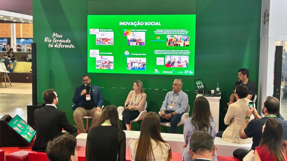 Esta fotografia captura um painel de debate em um ambiente de feira ou congresso, focado em temas de desenvolvimento social e inova&ccedil;&atilde;o.

Composi&ccedil;&atilde;o e Elementos Centrais
O Painel: Tr&ecirc;s palestrantes est&atilde;o sentados em poltronas brancas sobre um tablado. &Agrave; esquerda, um homem de barba e blazer azul fala ao microfone. Ao centro, uma mulher de blazer bege escuta atentamente. &Agrave; direita, um homem de camisa social azul clara completa o trio principal. Um quarto participante &eacute; parcialmente vis&iacute;vel no canto direito.

Plano de Fundo: O palco &eacute; emoldurado por uma grande parede verde. &Agrave; esquerda, est&aacute; escrita a frase "Meu Rio Grande t&aacute; diferente.". No centro da parede, uma tela digital exibe o t&iacute;tulo "INOVA&Ccedil;&Atilde;O SOCIAL" em letras brancas, cercado por seis quadros menores que apresentam fotos e nomes de projetos estrat&eacute;gicos, como "Bola pro Futuro" e "Olhar Adolescente".

Identifica&ccedil;&atilde;o Institucional: Na base da tela, aparecem logotipos de &oacute;rg&atilde;os como FAPERGS e o programa INOVA RS.

Perspectiva e Ambiente
O P&uacute;blico: A foto foi tirada da perspectiva da audi&ecirc;ncia. Em primeiro plano, vemos as costas e as cabe&ccedil;as de v&aacute;rias pessoas sentadas em bancos vermelhos e amarelos, observando a apresenta&ccedil;&atilde;o.

Atmosfera: O cen&aacute;rio &eacute; vibrante e profissional, t&iacute;pico de grandes eventos de tecnologia e gest&atilde;o p&uacute;blica, como a Gramado Summit mencionada no t&iacute;tulo do arquivo.

Contexto
A imagem ilustra a integra&ccedil;&atilde;o entre universidades, agenda clim&aacute;tica e inova&ccedil;&atilde;o, refor&ccedil;ando a apresenta&ccedil;&atilde;o de iniciativas de impacto social para a gest&atilde;o p&uacute;blica.