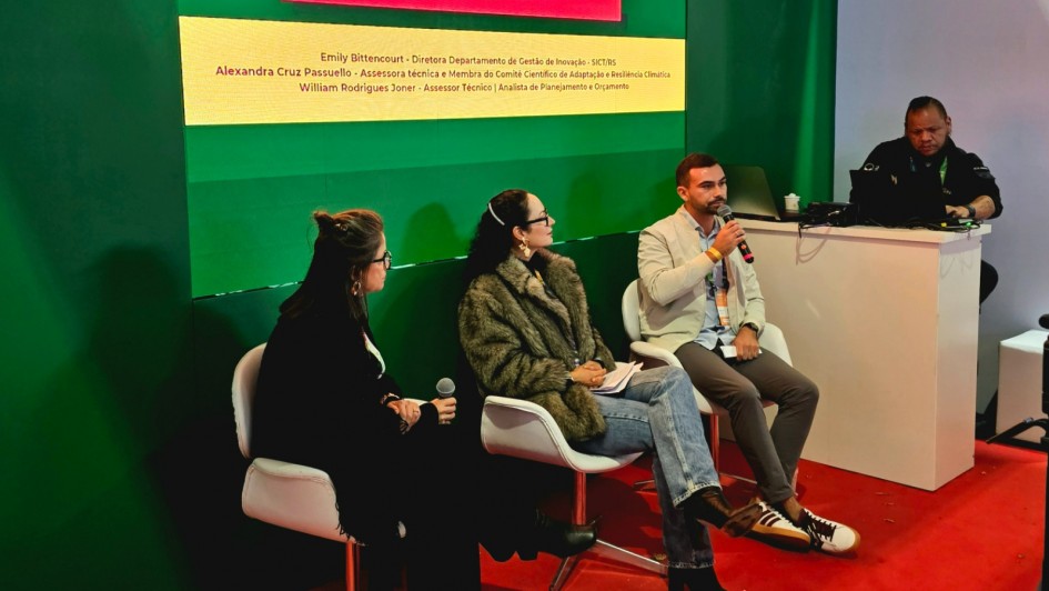 Tr&ecirc;s palestrantes est&atilde;o sentados em um palco, com um painel ao fundo. Um deles fala ao microfone. 