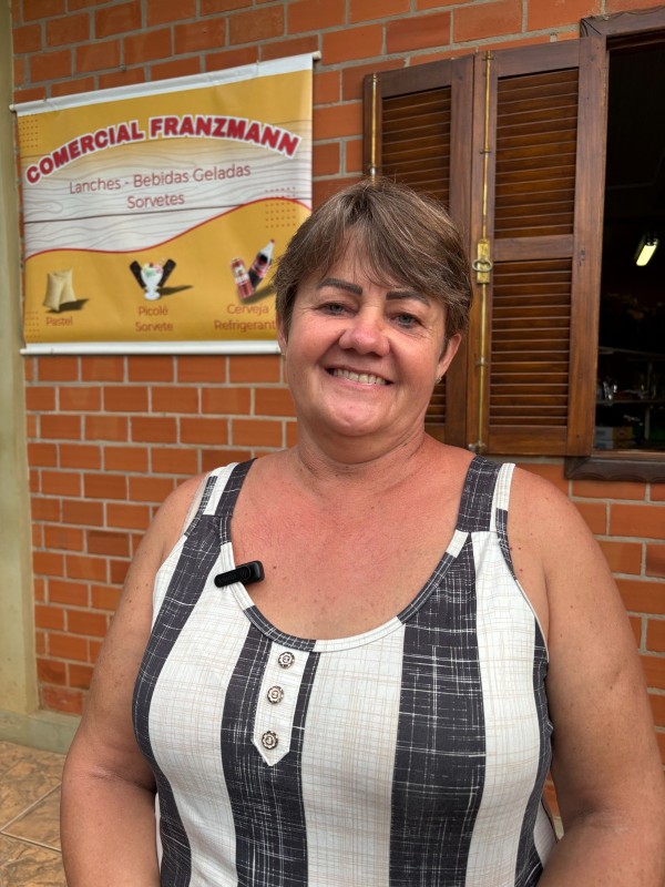 A imagem mostra uma mulher posicionada em frente a uma fachada de tijolos aparentes, em ambiente externo. Ao fundo, há uma janela de madeira com venezianas abertas, revelando o interior do estabelecimento. À esquerda, um banner fixado na parede traz o texto “COMERCIAL FRANZMANN”.