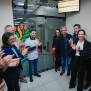 Cerim&ocirc;nia de inaugura&ccedil;&atilde;o em ambiente interno de unidade de sa&uacute;de, com grupo reunido diante de porta de vidro identificada como &ldquo;Sa&uacute;de 60+ RS &ndash; Servi&ccedil;o Especializado de Sa&uacute;de da Pessoa Idosa&rdquo;. Uma fita vermelha est&aacute; sendo cortada enquanto pessoas ao redor aplaudem.