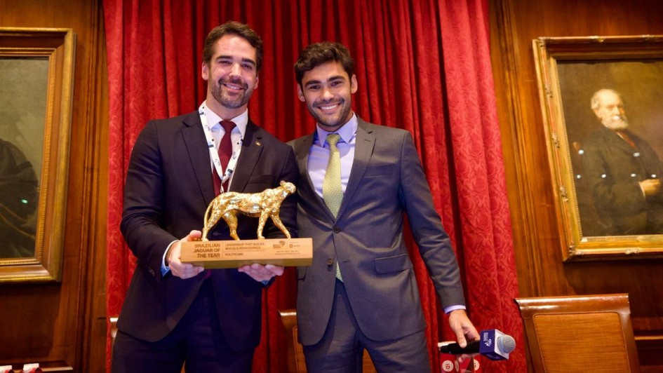 Esta &eacute; uma descri&ccedil;&atilde;o da imagem para fins de acessibilidade:

A fotografia mostra dois homens sorridentes posando para um registro formal em um ambiente interno luxuoso e cl&aacute;ssico.

&Agrave; esquerda: O governador Eduardo Leite, um homem branco de cabelos e barba aparados. Ele veste um terno azul-marinho, camisa branca e gravata bord&ocirc;. Ele segura com as duas m&atilde;os um trof&eacute;u que consiste em uma base de madeira retangular com uma escultura dourada de uma on&ccedil;a (jaguar) em cima. Na base, l&ecirc;-se o texto "BRAZILIAN JAGUAR OF THE YEAR".

&Agrave; direita: Um homem jovem, branco, de cabelos curtos e escuros. Ele veste um terno cinza, camisa lil&aacute;s clara e uma gravata em tom de verde amarelado. Ele est&aacute; com o bra&ccedil;o direito levemente apoiado nas costas do governador e segura um microfone com a m&atilde;o esquerda na altura da cintura.

Plano de fundo: Atr&aacute;s dos dois homens, h&aacute; uma grande e pesada cortina vermelha. Nas laterais, as paredes s&atilde;o revestidas com pain&eacute;is de madeira nobre (boiserie). &Agrave; esquerda e &agrave; direita, veem-se partes de quadros com molduras douradas cl&aacute;ssicas, retratando figuras hist&oacute;ricas.

Ambiente: A ilumina&ccedil;&atilde;o &eacute; quente e direcionada, destacando os sujeitos centrais. Na parte inferior, notam-se o topo de cadeiras de madeira e alguns objetos sobre uma mesa, como embalagens de &aacute;gua e outro microfone.

A imagem registra um momento de premia&ccedil;&atilde;o e reconhecimento internacional em um ambiente de prest&iacute;gio.